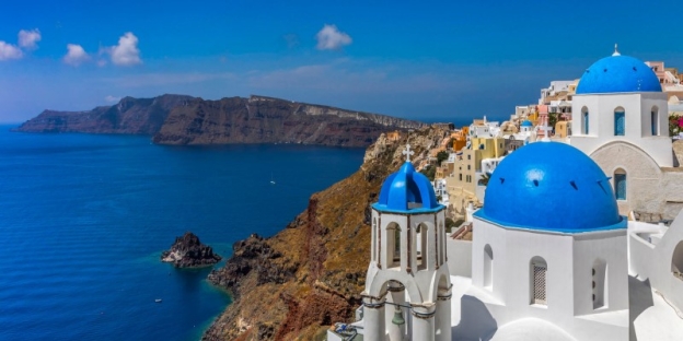 A white buildings with blue domes and a body of water with Santorini in the background AI-generated content may be incorrect.